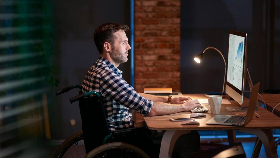 Ein Mann im Rollstuhl sitzt vor einem Arbeitstisch und tippt auf einer Tastatur.